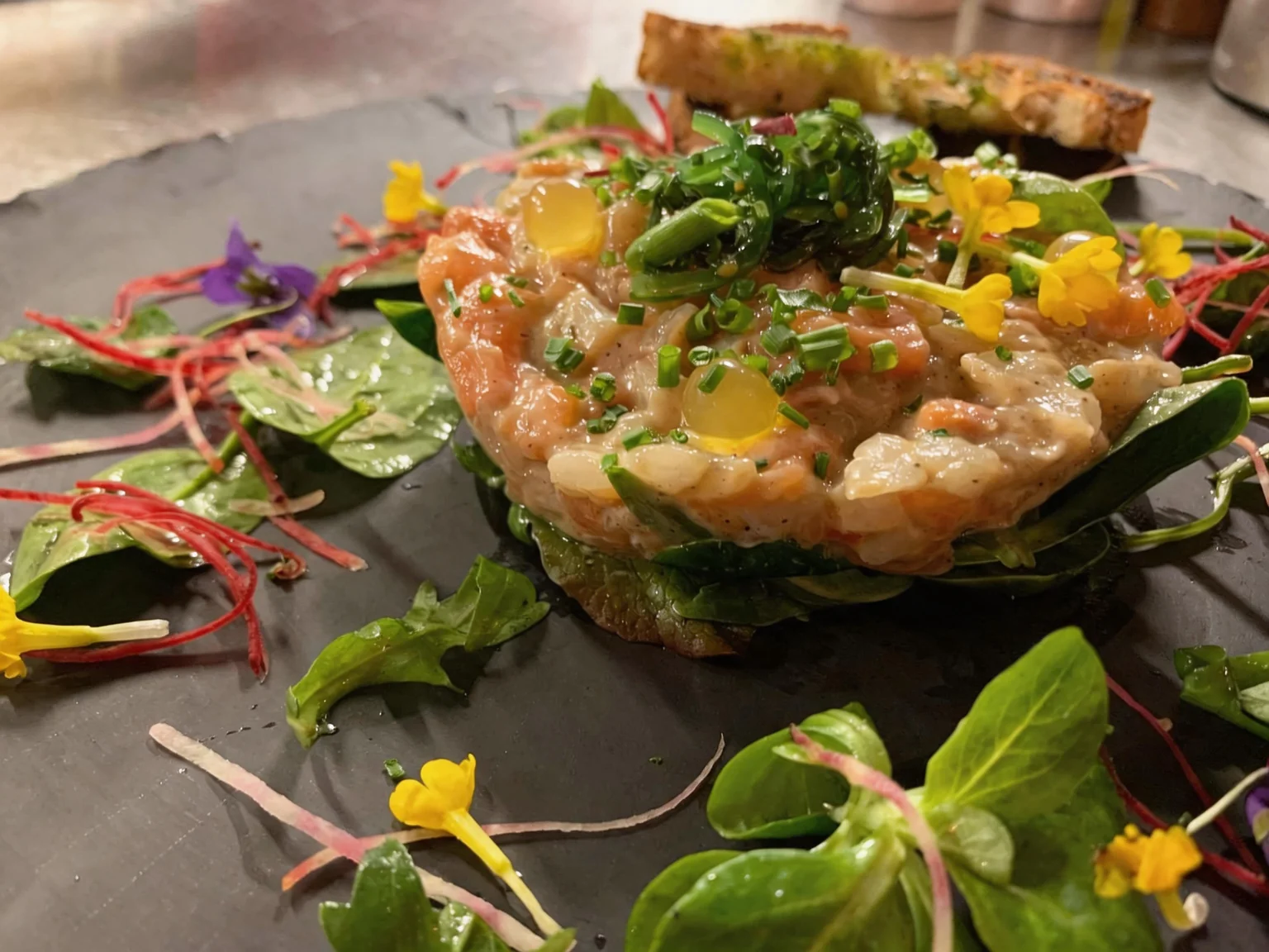 A tartare topped with herbs and flowers, served with a slice of grilled toast.