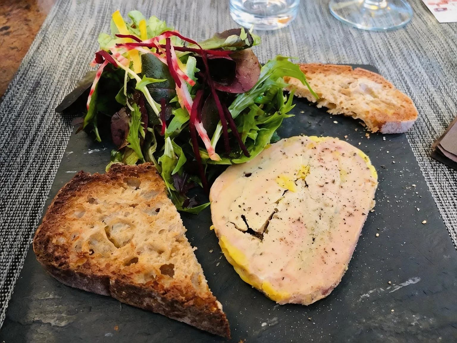 A slice of foie gras terrine, with a green salad and two slices of toasted bread, on a table with glasses.